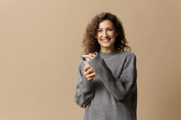 Happy curly beautiful lady in gray casual sweater chatting with boyfriend looks at camera posing isolated on beige pastel background. Social media, network, distance communication concept. Copy space