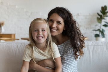 Young Hispanic stepmother sitting on couch embracing cute preschooler 5s daughter, having wide...