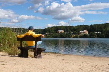 A young man in a yellow hoodie sits on a bench by the lake and looks into the distance. Man outdoors in early autumn.