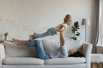 Young woman lying on sofa lifts on arms her preschooler daughter, play with 5s child at home, do acro yoga together in living room. Playtime with kids, happy motherhood, custody, family travel concept