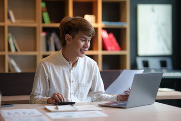 Fototapeta premium Young Asian businessman sit on his laptop reviewing his work happily and intently taking notes in the office.
