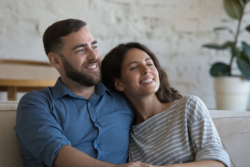 Happy loving couple relax on sofa smile looking into distance. Beautiful wife handsome husband daydreaming feel satisfied enjoy leisure at new own house. Homeowners family, relationships, love concept