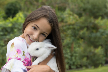 Smiling little girl is holding a white rabbit in her arms outdoors. Horizontally. 