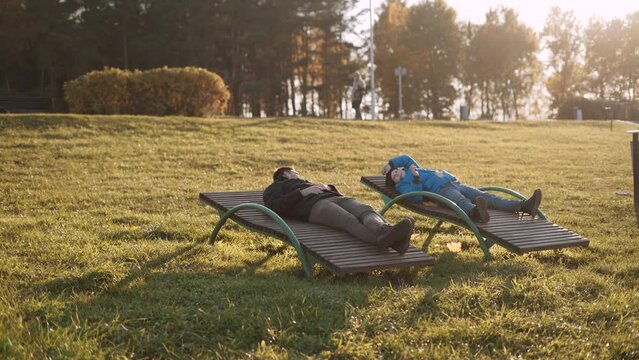 Family in warm casual clothes lie on deck chairs in sunny autumn park.