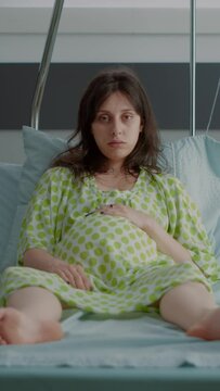 Vertical video: Portrait of pregnant person sitting in hospital ward bed with monitor preparing for child delivery at clinic. Caucasian young woman feeling impatient expecting labor contractions