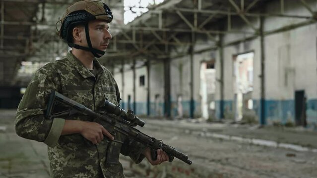 Fully equipped and armed caucasian soldier looking around damaged building during mission. Confident man wearing camouflage uniform walking trough large authorized factory with gun in hands.