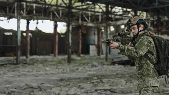 Professional female soldier with sniper rifle taking aim in destroyed industrial factory. Confident woman in uniform and helmet defending territory of ruined building.