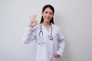 Happy smiling Asian female doctor wearing white medical uniform thumbs up ok with medical stethoscope on gray background medical concept.