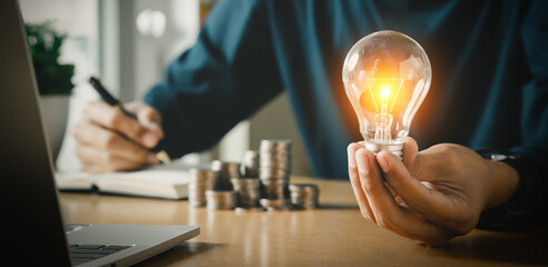 Businessman holding and putting lightbulb on coins stack on table for saving energy and saving money concept.