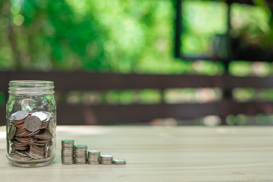 Picture of a clear bottle full of coins beside a stack of coins on table with a green backdrop of trees. saving money is an investment for lifestyle. Banking investment. financial and loan concept.