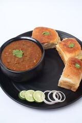Indian Pav bhaji, fast food dish from mumbai, vegetable curry served with a soft bread roll, served in a black plate with onion and lemon on white background