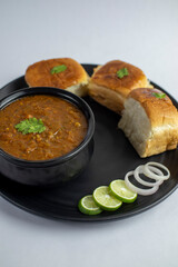 Indian Pav bhaji, fast food dish from mumbai, vegetable curry served with a soft bread roll, served in a black plate with onion and lemon on white background