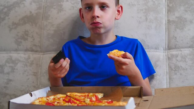 attractive caucasian boy 6-7 years old eats pizza from a box holding a tv remote and sitting on the couch. the child has a bite of pizza watching tv. kids love pizza for lunch