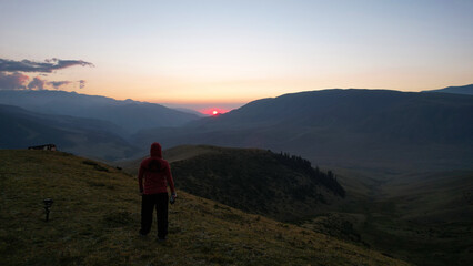 Fototapeta premium A guy in the mountains is watching the sunset. An epic orange-red sunset is reflected on small clouds and green hills. Bushes and forest grow in places on mountains. The guy is standing with his back