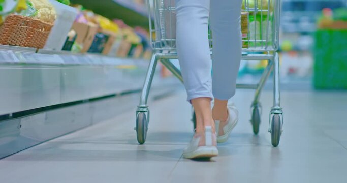 Women's feet in sneakers walking around the supermarket. Woman drives a grocery cart through a supermarket, rear view. 4k, ProRes