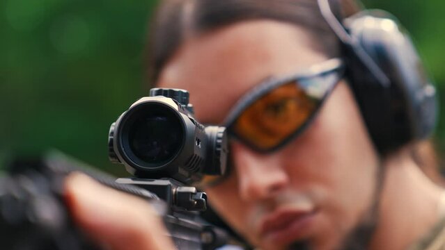 White man in safety goggles and headphones wearing camo t-shirt aiming submachine gun on firing range. Gun sight. Horizontal shot . High quality 4k footage