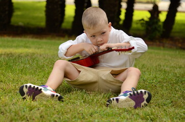 Little boy plays the acoustic guitar in the park. The child learns to play the uculele. Music...