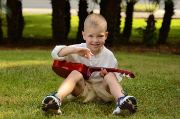Little boy plays the acoustic guitar in the park. The child learns to play the uculele. Music...