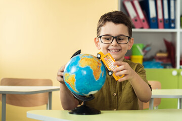 Cute caucasian schoolboy wearing glasses and sits at a desk in the classroom at elementary school. Little boy next the Globe playing with yellow school bus toy. Back to school concept