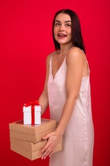 A young woman in an evening dress holds a lot of boxes with gifts for Christmas. A surprised model on a red background poses in the studio and looks into the camera