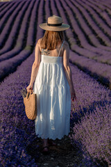 A middle-aged woman, blonde in a white dress and hat, walks through a lavender field with a basket