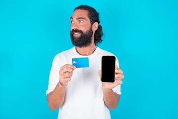 young bearded man wearing white T-shirt over blue studio background holding bank card modern device looking empty space