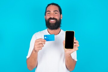 Photo of adorable young bearded man wearing white T-shirt over blue studio background holding credit card and Smartphone. Reserved for online purchases