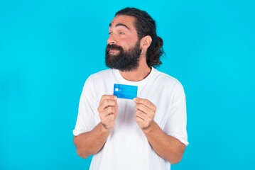 Photo of cheerful young bearded man wearing white T-shirt over blue studio background hold debit card look empty space