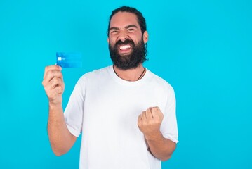 Fototapeta premium Excited happy positive cheerful smiling young bearded man wearing white T-shirt over blue studio background hold credit card raise fist in victory