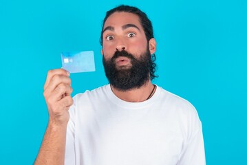 Photo of young bearded man wearing white T-shirt over blue studio background amazed shocked hold credit card payment