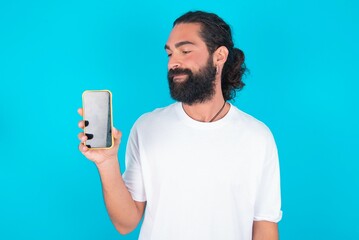 Photo of interested young bearded man wearing white T-shirt over blue studio background hold telephone look side empty space screen