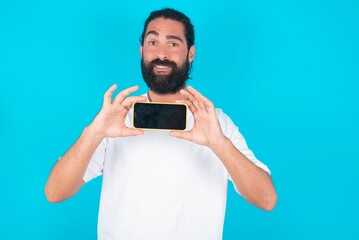 Cheerful cheery content young bearded man wearing white T-shirt over blue studio background holding in hands device hobby smm post blog