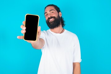 Charming adorable young bearded man wearing white T-shirt over blue studio background holding modern device, showing black screen smartphone