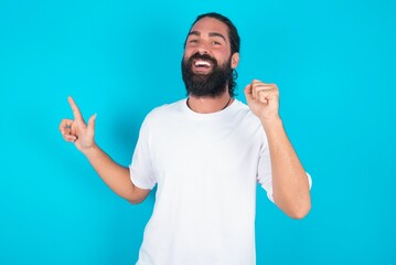 young bearded man wearing white T-shirt over blue studio background points at empty space holding fist up, winner gesture.