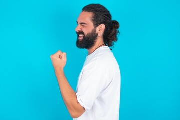 Portrait of funny young bearded man wearing white T-shirt over blue studio background shout yeah...