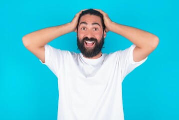 Fototapeta na wymiar Cheerful overjoyed young bearded man wearing white T-shirt over blue studio background reacts rising hands over head after receiving great news.