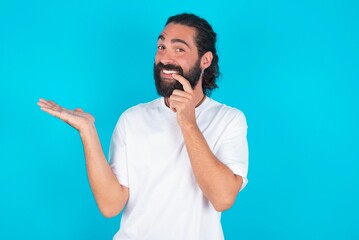 Positive young bearded man wearing white T-shirt over blue studio background advert promo touch finger teeth