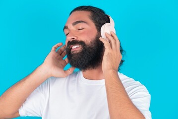 Fototapeta premium young bearded man wearing white T-shirt over blue studio background with headphones on her head, listens to music, enjoying favourite song with closed eyes, holding hands on headset.