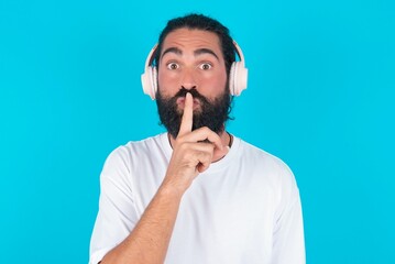 young bearded man wearing white T-shirt over blue studio background making hush gesture with finger on her lips wearing  wireless headphones. Be quiet.