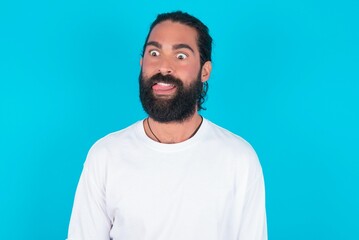 Funny young bearded man wearing white T-shirt over blue studio background makes grimace and crosses...
