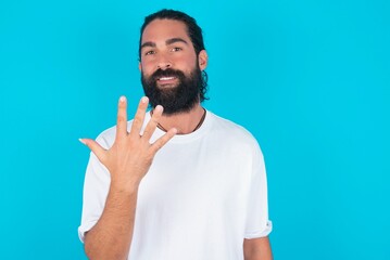 Naklejka premium young bearded man wearing white T-shirt over blue studio background smiling and looking friendly, showing number five or fifth with hand forward, counting down