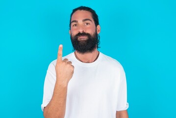 Naklejka premium young bearded man wearing white T-shirt over blue studio background smiling and looking friendly, showing number one or first with hand forward, counting down