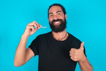 young bearded man wearing black T-shirt over blue studio background holding an invisible braces aligner and rising thumb up, recommending this new treatment. Dental healthcare concept.