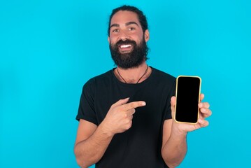 Attractive cheerful young bearded man wearing black T-shirt over blue studio background holding in hands cell showing black screen