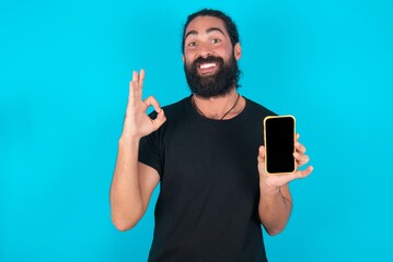 young bearded man wearing black T-shirt over blue studio background holding in hands cell showing...