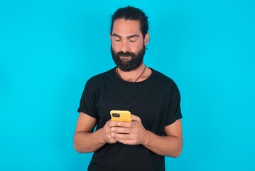 Excited young bearded man wearing black T-shirt over blue studio background winking and eye hold smart phone use read social network news