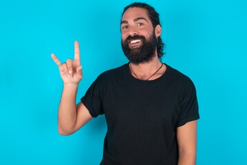 young bearded man wearing black T-shirt over blue studio background doing a rock gesture and smiling to the camera. Ready to go to her favorite band concert.