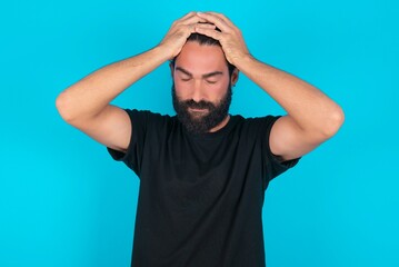 young bearded man wearing black T-shirt over blue studio background suffering from strong headache...