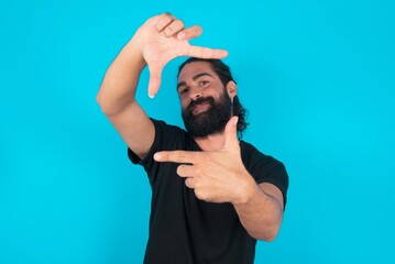 young bearded man wearing black T-shirt over blue studio background making finger frame with hands. Creativity and photography concept.
