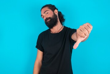 young bearded man wearing black T-shirt over blue studio background feeling angry, annoyed, disappointed or displeased, showing thumbs down with a serious look